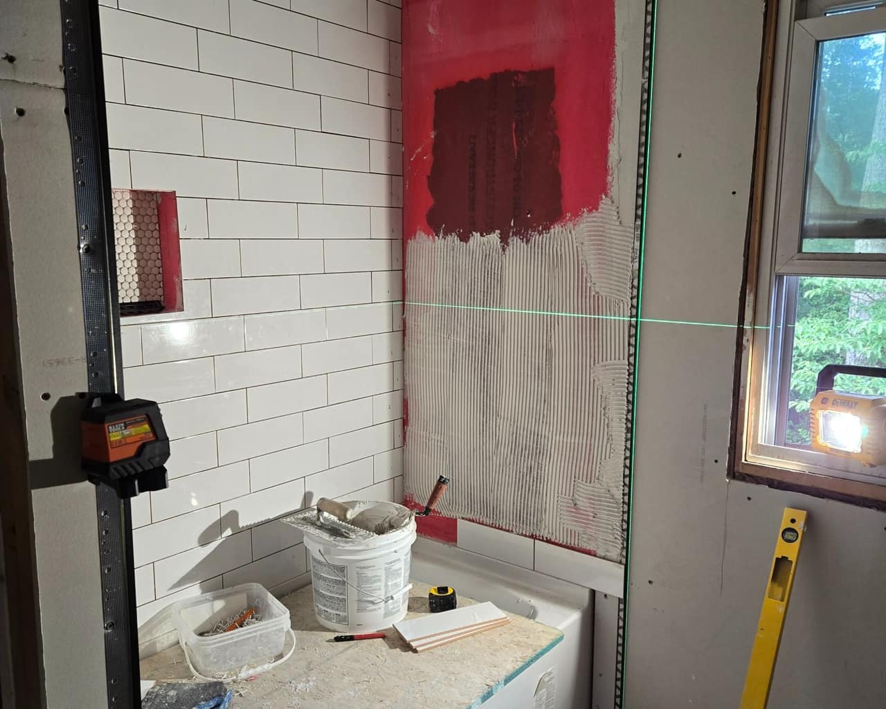 White subway tile being set over red waterproofing membrane and combed thinset, with a green laser line running across the wall